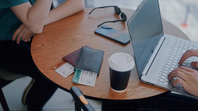 Two travelers sitting at a cafe table with laptop, passports, and boarding passes in an airport