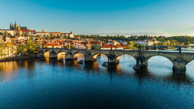 Aerial view of the historic Charles Bridge and Prague Castle overlooking the Vltava River with red-roofed buildings under a clear blue sky in Prague, Czechia.