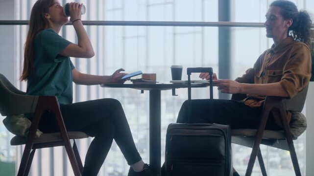 Two travelers, a couple, share a table with a laptop, two coffee cups, and their suitcases parked beside them. The scene captures the modern reality of waiting for a flight while staying connected
