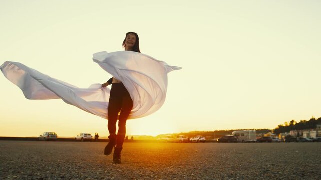 Woman dances in the sunset with flowing fabric at a beach location during golden hour time