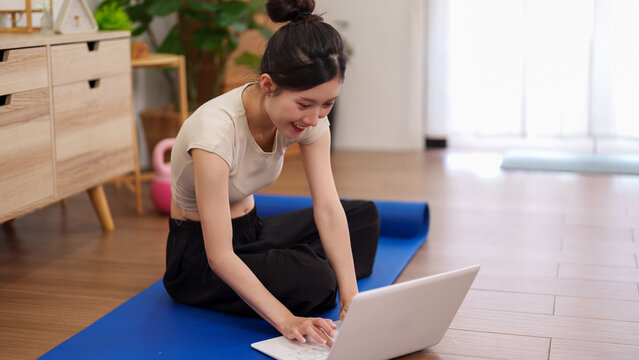 Asian women fitness instructor using laptop computer following online yoga workout tutorial at home exercise on yoga mat smiling cheerful healthy lifestyle