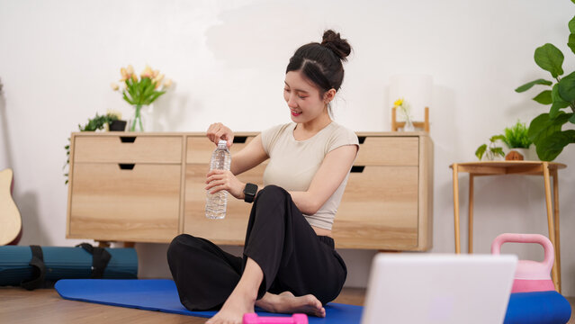 Asian woman sitting on yoga mat holding water bottle after home workout fitness exercise training session in modern living room with plants healthy lifestyle