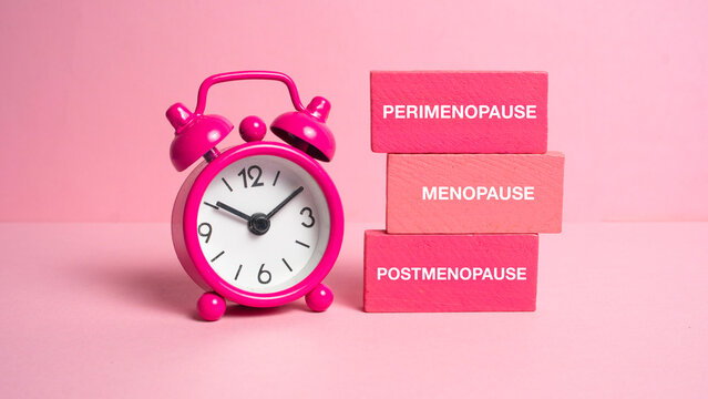 Pink alarm clock next to wooden blocks labeled with perimenopause menopause and postmenopause stages representing the biological clock.