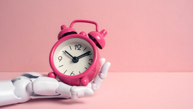 Description: White robotic hand holding a pink vintage alarm clock against a pastel pink background representing AI time management and future technology.