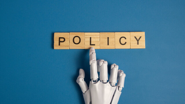White robotic hand pointing to the word policy on wooden blocks over a blue background representing artificial intelligence ethics and government regulations.