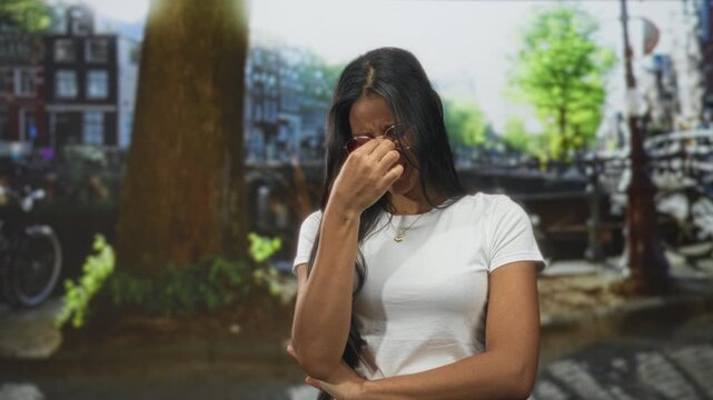 Woman pinches nose with fingers, wearing glasses and white t shirt, standing by canal and bicycle on street; disgust.
