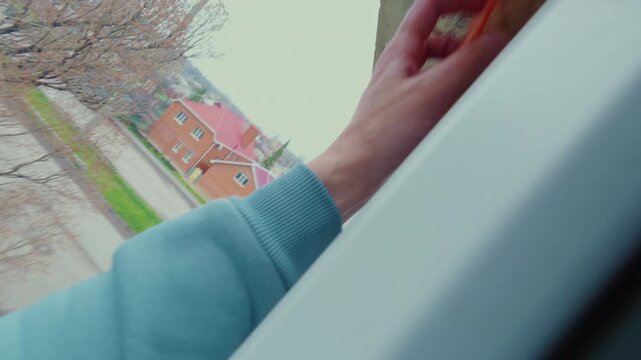 A man is sponging the glass of a plastic window outside at the height of the fourth floor. The process of washing dirty windows of a residential apartment. From the first person.