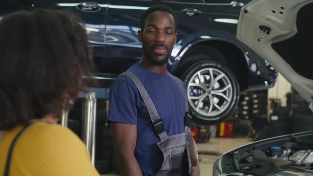 Car mechanic and a female client shaking hands after a vehicle repair, the repairman closing the hood and returning her car keys.