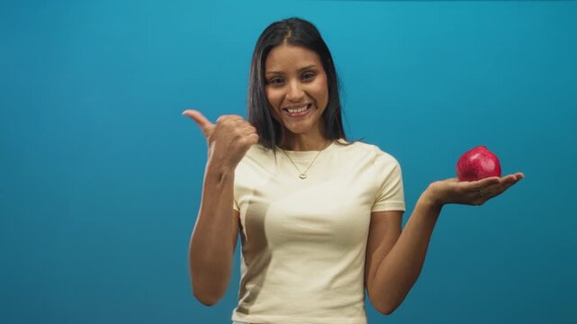 Woman holding pomegranate in open palm while smiling and pointing thumb toward herself in studio; healthy choice cheerful.