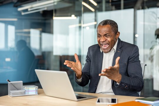 Stressed mature professional man throwing hands up in exasperation and shouting at his laptop, experiencing a computer problem or work pressure during an intense business day in the office