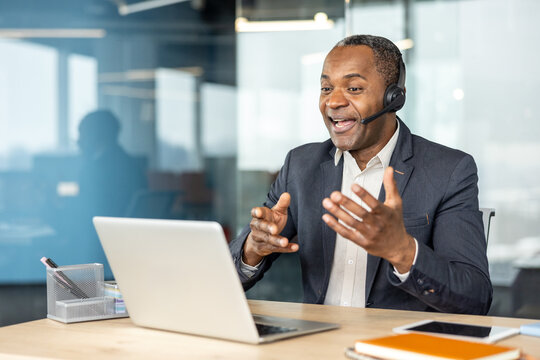 Mature businessman with headset talking and gesturing during a laptop video call in a modern corporate office, representing remote communication, collaboration and professional interaction