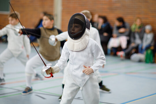 Little kid boy fencing on a fence competition. Child in white fencer uniform with mask and sabre. Active kid training with teacher and children. Healthy sports and leisure.