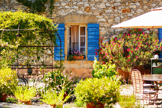 Sunny courtyard in Provence France with stone house, blue shutters and blooming flowers, cozy garden terrace, charming rural scene.