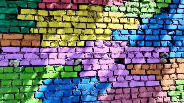 A sunlit, multi-colored brick wall painted in bold blocks of yellow, blue, pink, green, and purple stretches diagonally, with rough textures and uneven stones adding depth and character.