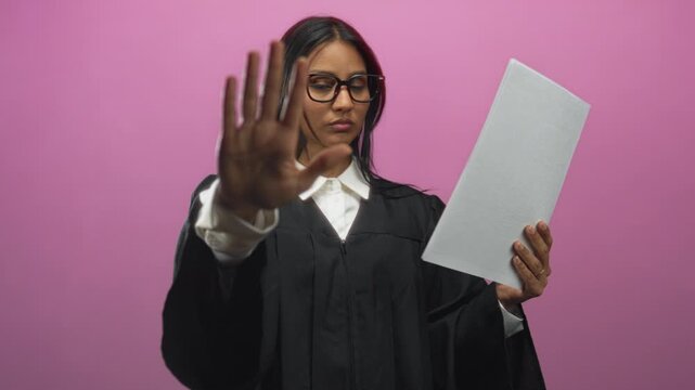 Woman judge in black robe holding document with hand raised palm forward stop gesture in a studio; authority duty.