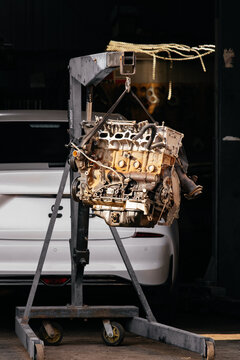 Car engine removed on a hydraulic crane in an auto repair shop. Process of major engine overhaul or replacement in a workshop, white car visible in the background.