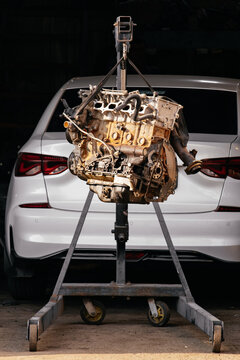 Car engine removed on a hydraulic crane in an auto repair shop. Process of major engine overhaul or replacement in a workshop, white car visible in the background.