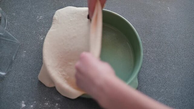 Hands are seen shaping dough to fit a pie dish in a kitchen setting. The action involves lifting and placing the dough with care to create a crust