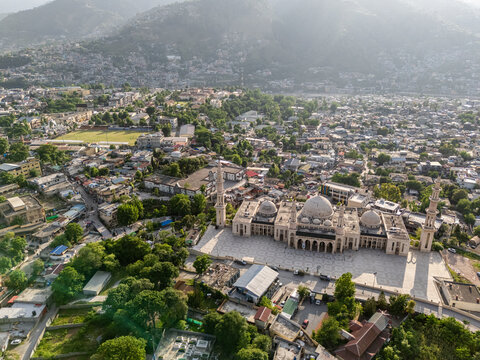 Aerial view of the grand Jalalabad Mosque with its white domes and minarets surrounded by the dense urban landscape and misty mountains of Muzaffarabad, Azad Kashmir, Pakistan.