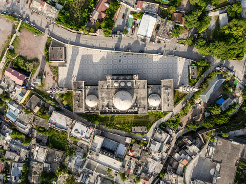 Aerial view of the grand mosque featuring white domes and a large patterned courtyard surrounded by urban buildings and lush greenery in Muzaffarabad, Azad Kashmir, Pakistan.