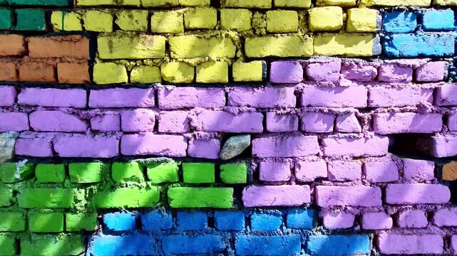 A close-up of a vividly painted brick wall showcases uneven rows of green, blue, yellow, orange, and lavender blocks, with rough mortar, embedded stones, and dappled sunlight enhancing its textured.