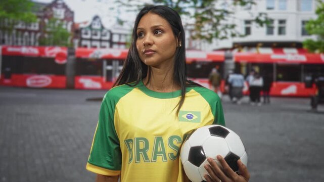 Woman in yellow brazil football jersey holds a soccer ball and wipes her face with one hand while standing on a crowded city street; disappointment.