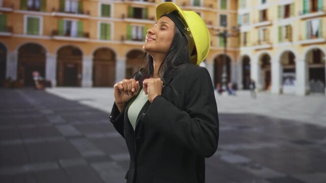 Woman wearing yellow hardhat smiles with eyes closed and tilts head back on street plaza with arched colonnades, blazer on; confidence progress.