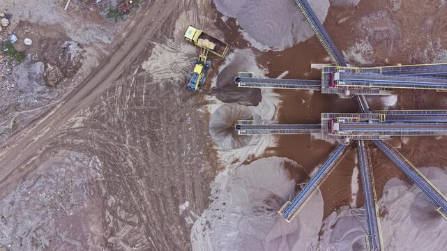 Large machinery is moving materials at an excavation site. Conveyors distribute sand and gravel while trucks deliver and collect resources for construction activities.