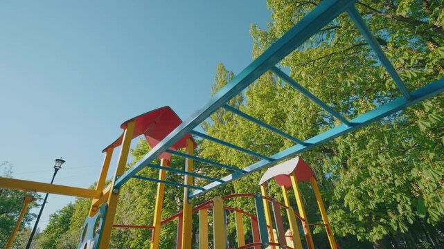 Colorful Monkey Bars on Children Playground &mdash; Bottom-Up Wide-Angle View | Blue Yellow Red Metal Overhead Ladder Against Clear Blue Sky and Green Chestnut Trees | Outdoor Kids Equipment