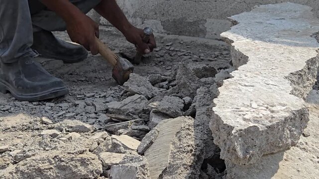 A worker uses a sledgehammer and chisel to manually break a reinforced concrete slab. The ground is covered in debris, rubble, and gravel from the demolition process, showing intense physical labor.