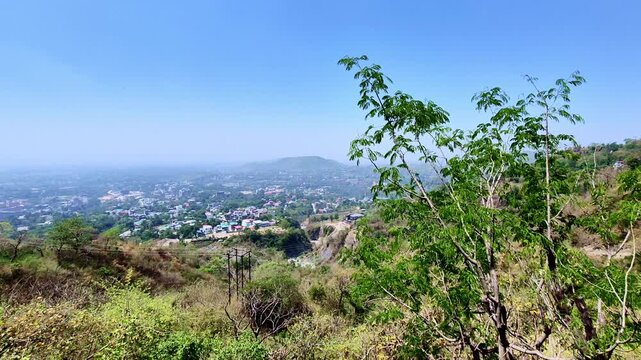 A sweeping daytime view from Mussoorie overlooks a hazy valley dotted with houses and greenery, framed by sparse hillside vegetation and a lone tree against a clear blue sky.