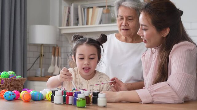 Asian little girl painting colorful easter eggs with brush while grandmother helping her at kitchen table, Three generations family spring holiday celebration, creative art craft learning, easter day