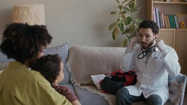 Handheld medium long shot of male multiethnic paediatrician preparing stethoscope while examining breath of small sick patient sitting on mothers lap during home appointment