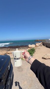A picturesque view of La Caleta Beach in Cadiz, Spain, featuring a black SUV and a Bialetti moka pot against the backdrop of the ocean