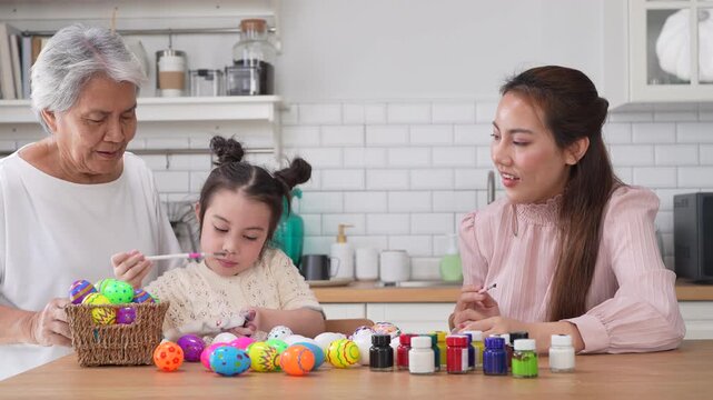 Asian little girl painting colorful easter eggs with brush while grandmother helping her at kitchen table, Three generations family spring holiday celebration, creative art craft learning, easter day
