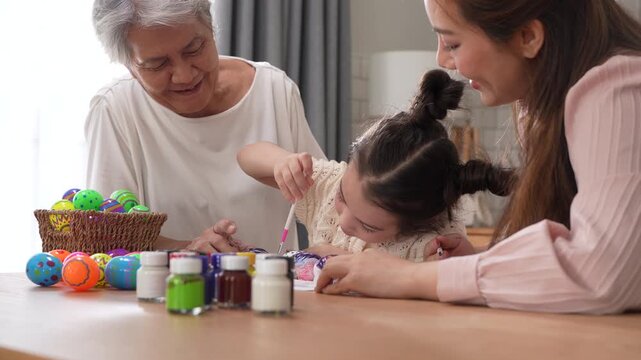 Asian little girl painting colorful easter eggs with brush while grandmother helping her at kitchen table, Three generations family spring holiday celebration, creative art craft learning, easter day