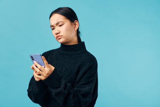 Woman in black sweater looking confused or frustrated at smartphone with blue background. Expression of doubt or concern while checking mobile device in casual studio setting.