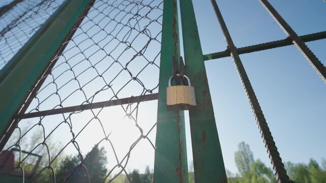 Locked Chain-Link Gate Closed with Padlock &mdash; Bottom-Up Wide-Angle View | No Entry Restricted Access Concept with Sun Flare Backlight | Rusty Metal Fence Against Blue Sky