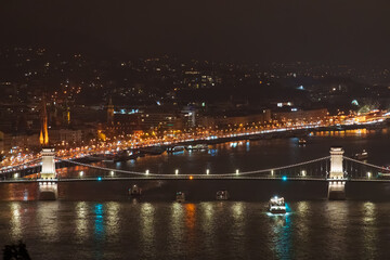 Naklejka premium Night View of Széchenyi Chain Bridge over Danube River in Budapest, Hungary; Illuminated Suspension Bridge with City Lights and Boat Traffic on Water at Nighttime