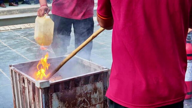 A metal burn bin filled with blazing debris emits thick white smoke and bright orange flames, as a nozzle sprays water onto the fire, burst of steam and scattering droplets across the tiled ground.