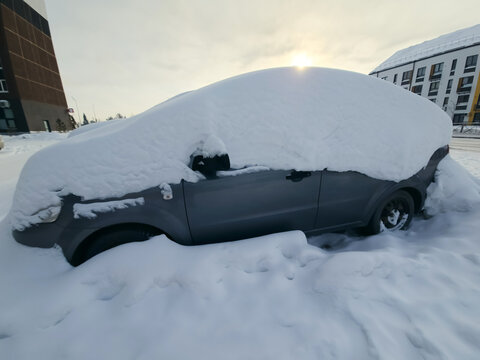 Snow covers the roofs and windshields of parked cars in a residential area. The sun sets in the background