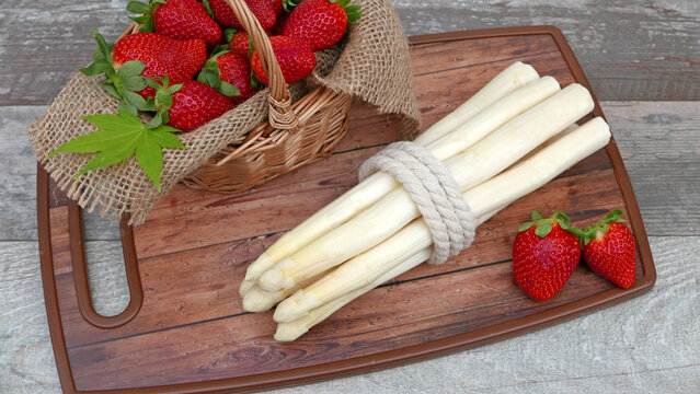 A wooden board with bundles of white asparagus and a basket full of strawberries.