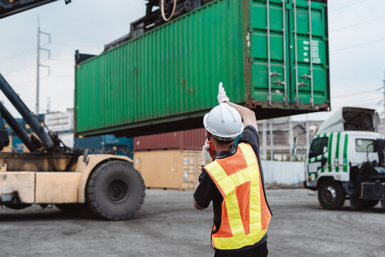 Logistics worker directing reach stacker lifting green container. Male operator using walkie-talkie for cargo transport and heavy equipment management.