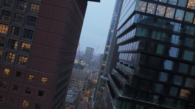 Dutch angle aerial view moving down between modern skyscrapers with illuminated office windows. Busy city street below with traffic at twilight