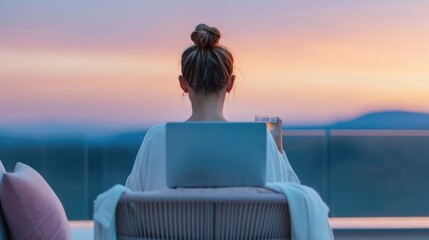 Obraz premium View from behind of a woman relaxing on a balcony with her laptop and a drink enjoying a vibrant sunset