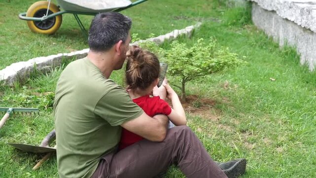 A father and his little girl take a break from gardening to look at a phone together, blending family activities with technology.
