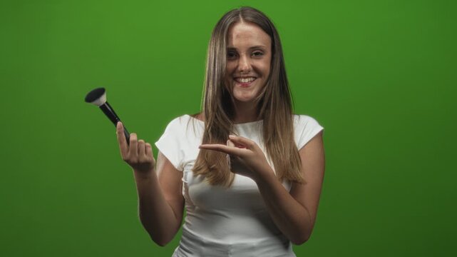 Woman holding makeup brush and pointing finger in studio green screen while smiling and presenting the brush; playful confidence.