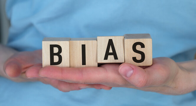 Hands gently hold wooden blocks spelling BIAS. Soft blue shirt adds calm, neutral backdrop.