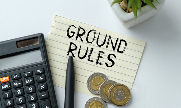 Black pen rests beside calculator and coins. Note reads GROUND RULES in bold, handwritten script. White table holds small plant in background.