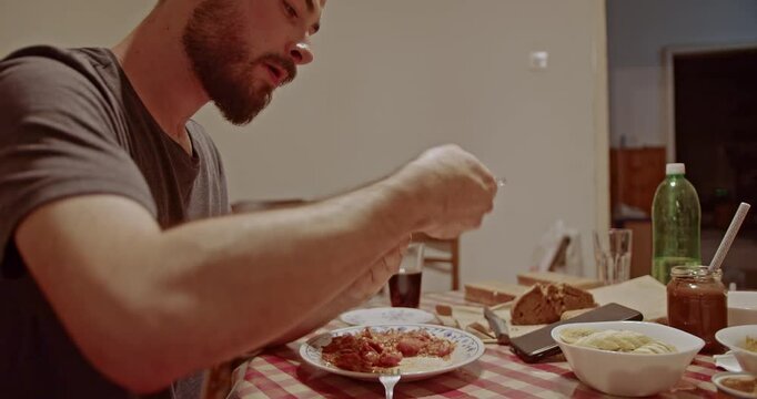 A bearded man savors a simple, homemade meal at a rustic dining table. He carefully spreads food onto a piece of bread, enjoying his personal and relaxed dining experience at home.